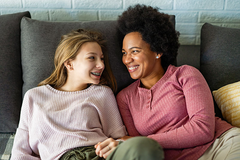 January 2024 Charity of the Month: Foster Hope. Image: A middle-aged black woman and a teen white girl sit together on a sofa, looking at each other and laughing. 