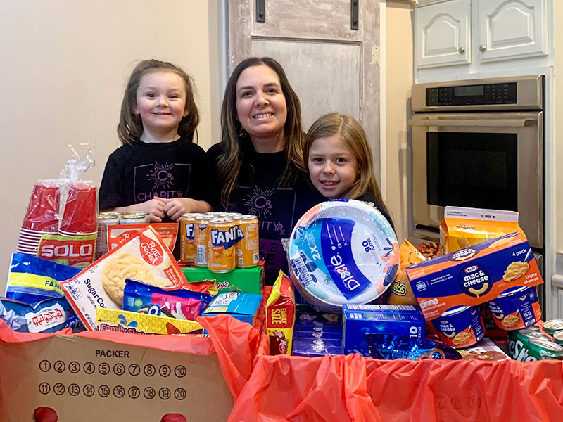 Image of a woman and two young girls standing behind two boxes full of food to be donated. CharityRx Spreading Hope Through Donations
