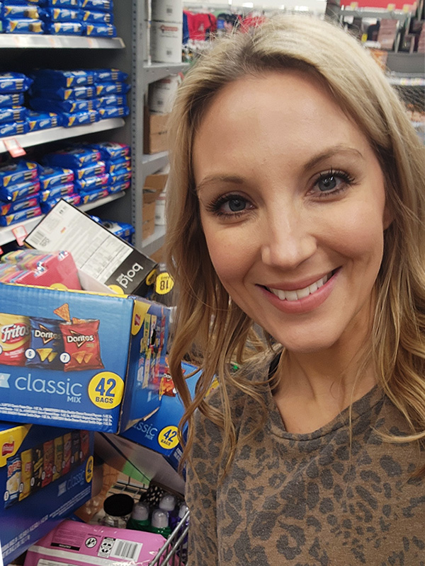 Image of a smiling blonde woman standing in front of a grocery cart full of snacks and personal care items. CharityRx Supports Women and Children