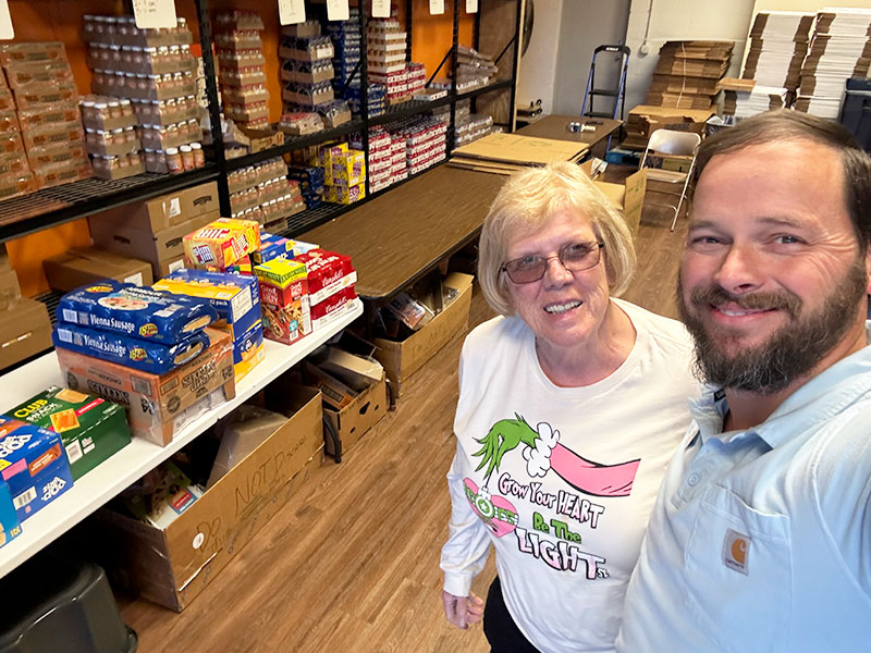 Image of a man and woman inside a warehouse or storage room next to shelves and a table stacked with boxes and cans of food. CharityRx Spreading Hope Through Donations