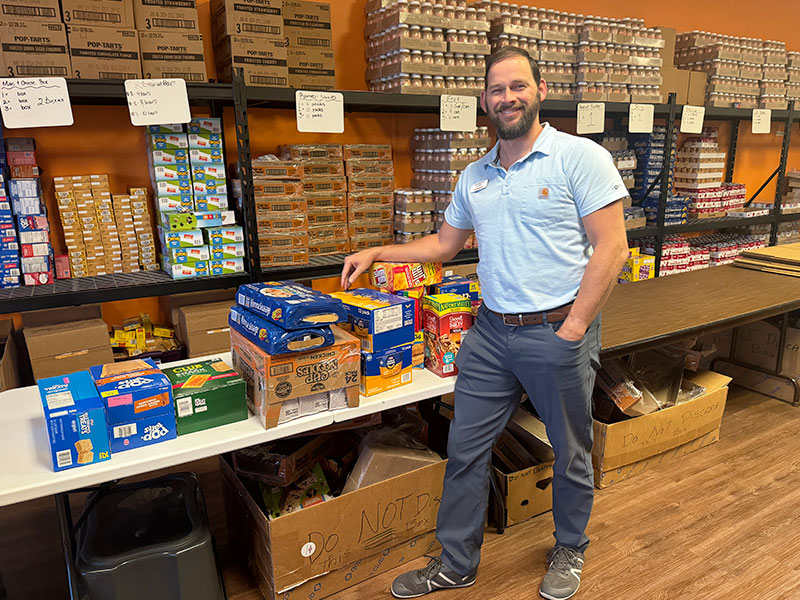 Image of a man standing next to a table with boxes food stacked on it inside a warehouse or storage room.
