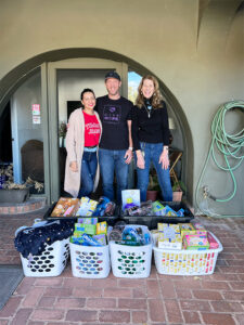 Image of three adults standing outside a building behind six large baskets full of donated clothing, feminine hygiene products, and housewares. CharityRx Supports Women and Children