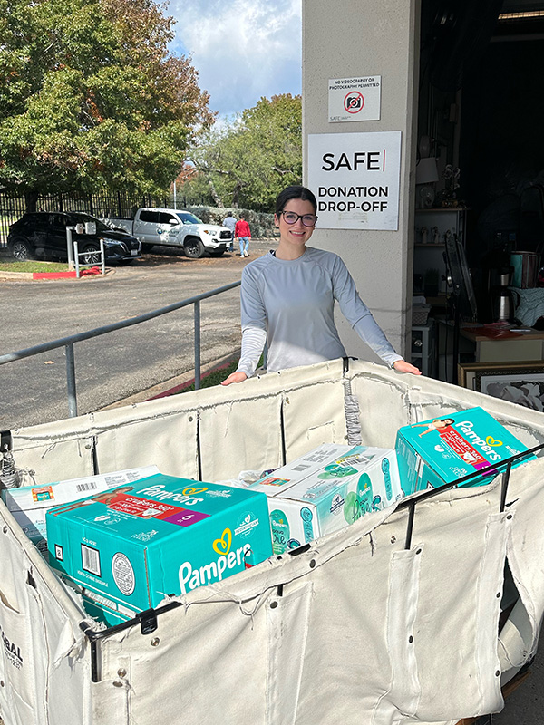 Image of a woman standing behind a wheeled bin full of donated diapers, laundry detergent, and board games. CharityRx Supports Women and Children