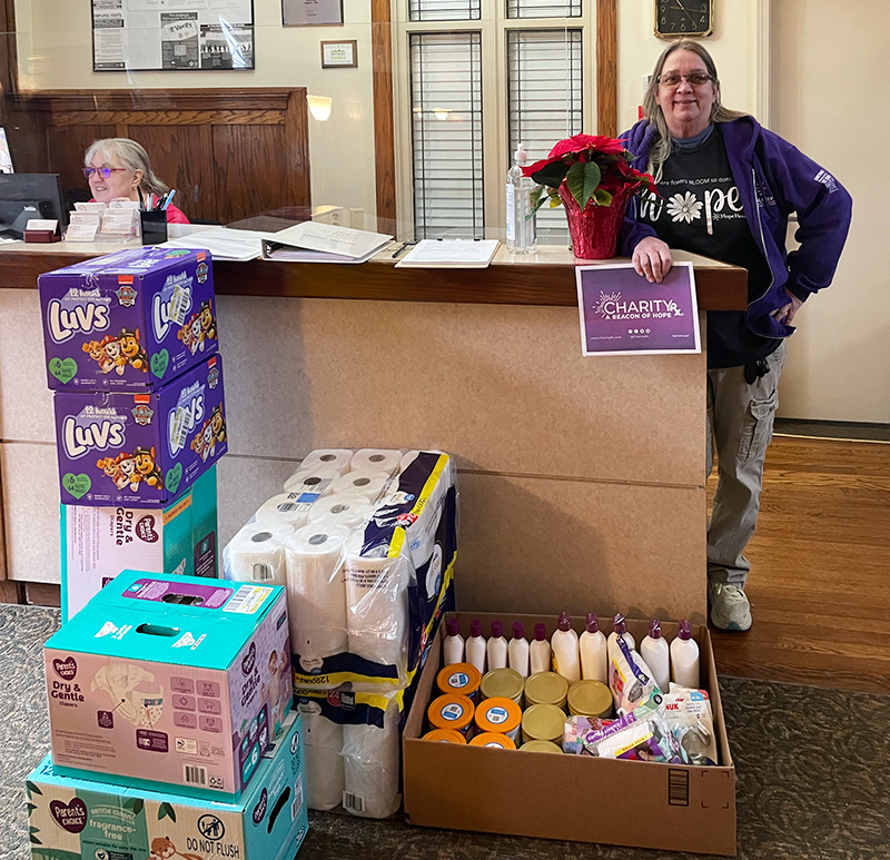 Image of two women behind the counter at Hope House of Kansas City. One woman holds a CharityRx sign and stands behind a stack of baby formula, diapers, and paper towels. CharityRx Supports Women and Children