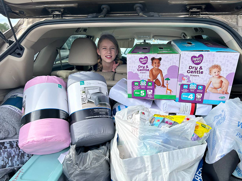 Image of the back of a vehicle with the hatch open and packed full of items to be donated. A smiling young girl peeks over the back seat and the pile of items. CharityRx Supports Women and Children