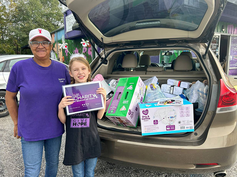 A woman and a young girl stand next to the open hatchback of a car full of diapers, wipes, sheets, and other supplies. The girl is wearing a CharityRx shirt and holding a CharityRx sign. The supplies were donated to a women's shelter in Florida.