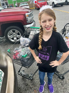 Image of a young blonde girl with braided hair winking and smiling at the camera. She stands in front of two shopping carts full of items to be donated. CharityRx Supports Women and Children