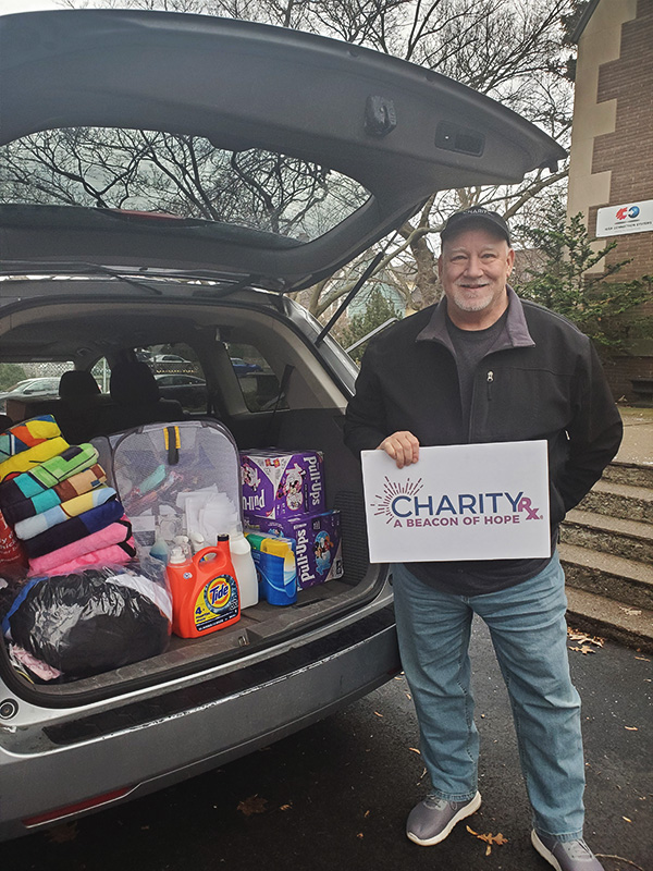 Image of a smiling man holding a sign that reads: CharityRx, a beacon of hope. He stands behind a vehicle fill of items to be donated. CharityRx Supports Women and Children