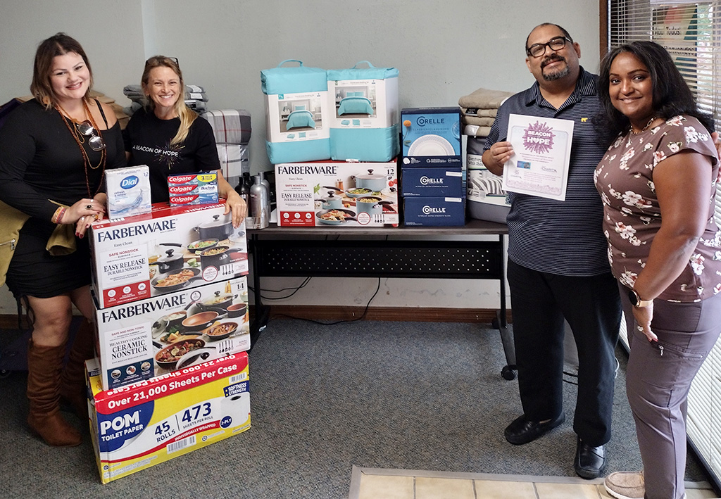 Image of three women and a man next to a table stacked with donated  bedding, dishes, pot and pan sets, and household items. The man is holding a Beacon of Hope certificate. CharityRx Supports Women and Children