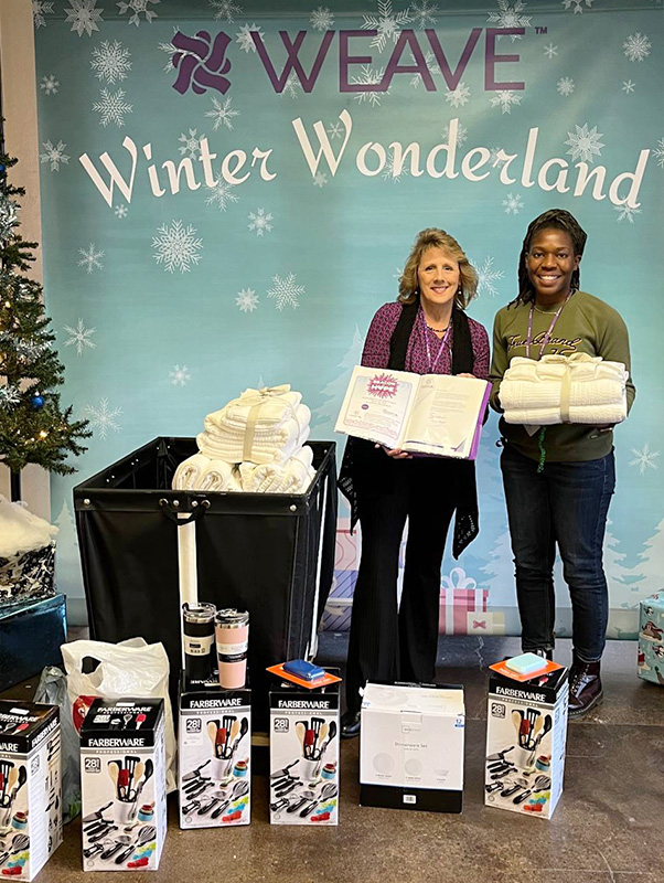Image of two women standing in front of a winter backdrop that reads, "Weave Winter Wonderland." The women stand behind a bin full of donated towel sets, kitchen utensils, power banks, and insulated cups. CharityRx Supports Women and Children