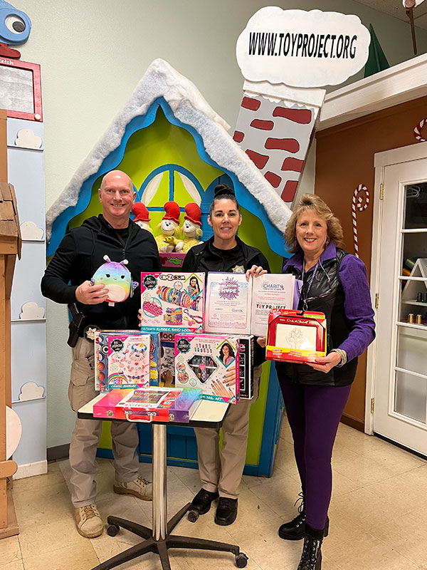 Image of a man and two women standing next to a stack of donated toys and gifts. CharityRx Spreading Hope Through Donations