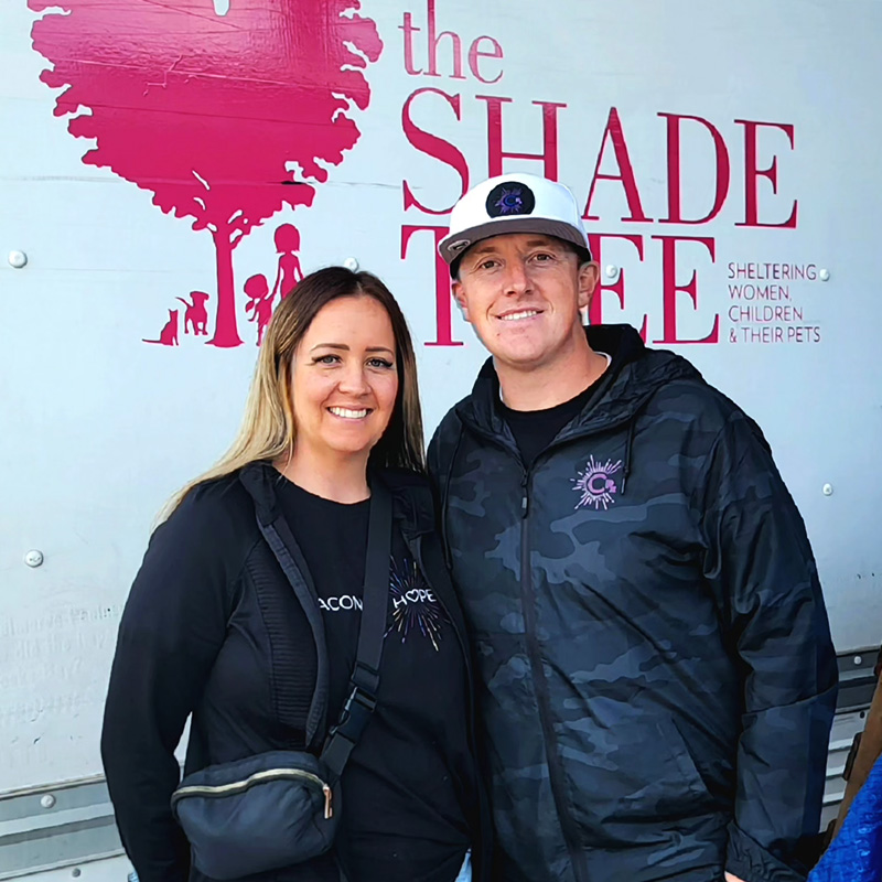 Image of a smiling man and woman standing in front of a sign that reads, "The Shade Tree." CharityRx Supports Women and Children