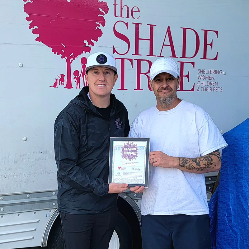 Image of two men holding a Beacon of Hope certificate and standing together in front of a sign that reads, "The Shade Tree." CharityRx Supports Women and Children