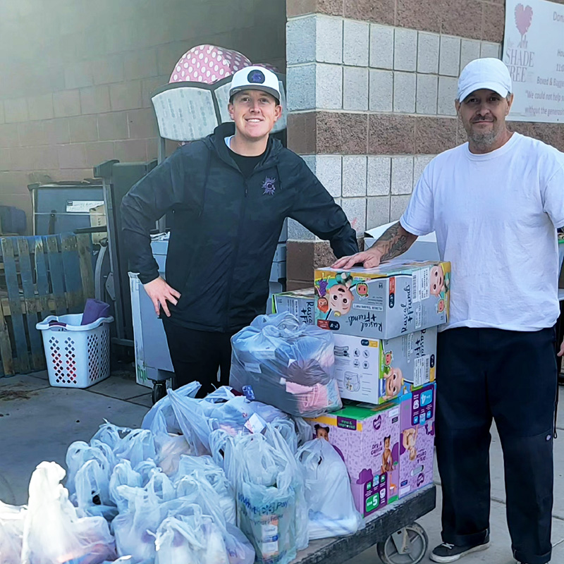 Image of two smiling men standing next to a cart loaded with donated feminine hygiene items, diapers, and personal care items. CharityRx Supports Women and Children