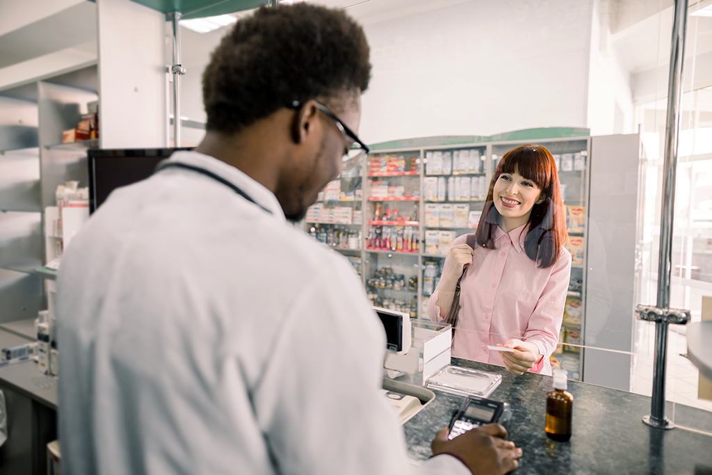 Image of a smiling woman handing a card to the pharmacist behind a glass window. 