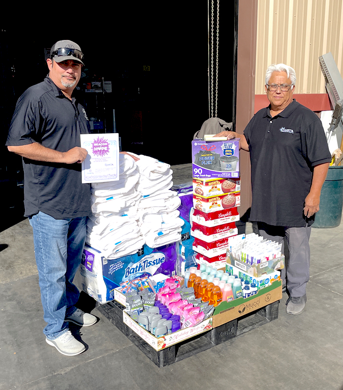 Image of two men standing next to a pallet of donated towels, paper towels, caned food, and personal care items. CharityRx Supports Women and Children
