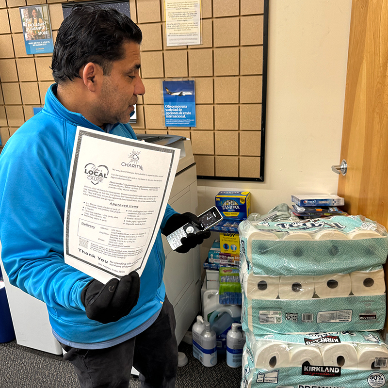 Image of a man holding a list of requested items from the Courageous Women Association Shelter and standing next to a stack of donated paper towels, hair care products, and other items. CharityRx Supports Women and Children