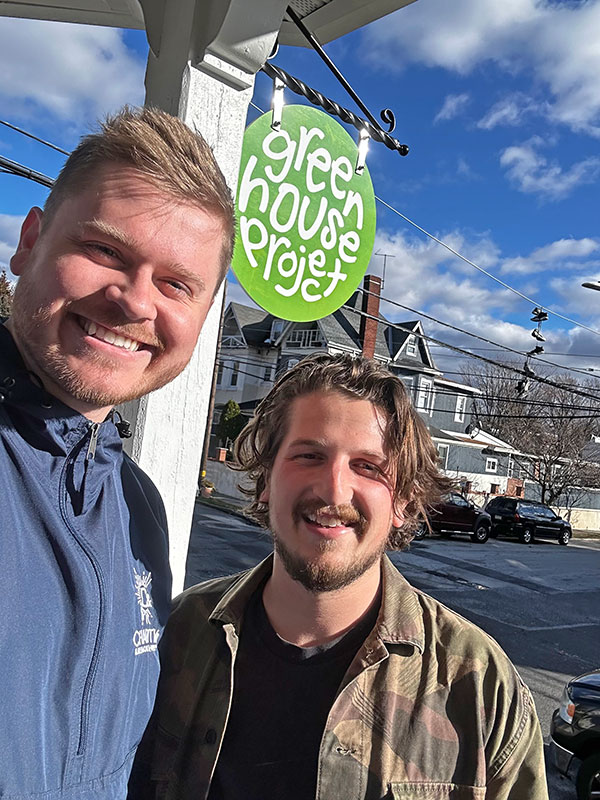 Image of two men smiling into the camera with the Greenhouse Project sign in the background.