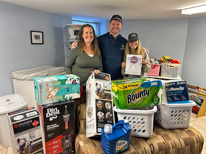 Image of a man and two women standing inside a house behind a donated vacuum and two laundry baskets full of household items, cleaning supplies, and diapers. CharityRx Supports Women and Children