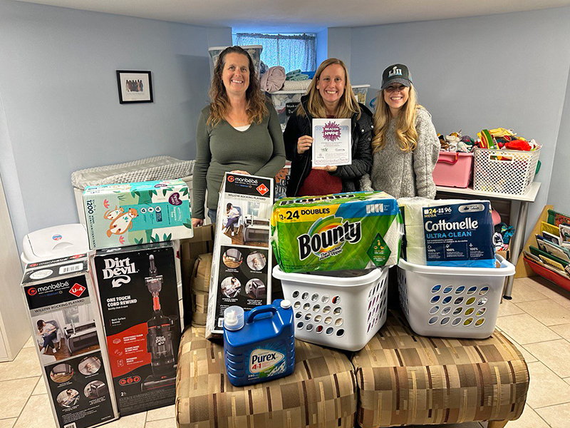 Image of three women standing inside a house behind a donated vacuum and two laundry baskets full of household items, cleaning supplies, and diapers. CharityRx Supports Women and Children
