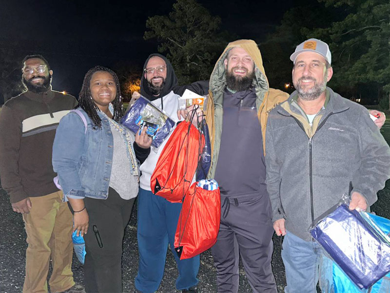 Image of four men and a woman standing in a line at night. Each person is holding a backpack or bag of supplies. CharityRx Spreading Hope Through Donations