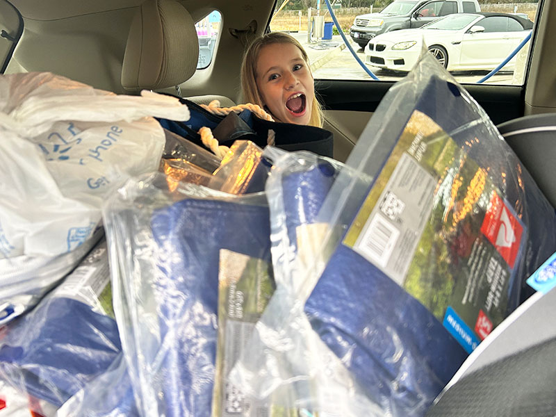 Image of an excited young girl in the back seat of a car mostly hidden by tarps and packages for donation.
