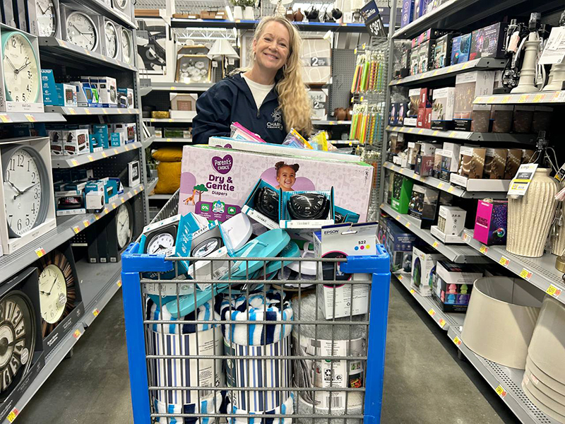 Image of a smiling blonde woman pushing a shopping cart full of diapers, blankets, and household items. CharityRx Supports Women and Children