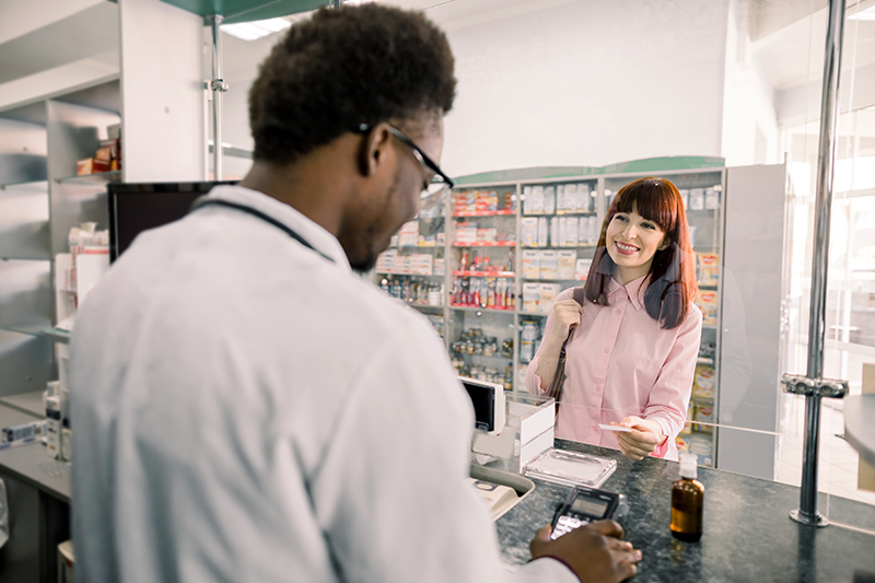 Image of a smiling, red-haired woman handing the CharityRx Prescription Discount Card to a young Black pharmacist behind the counter.