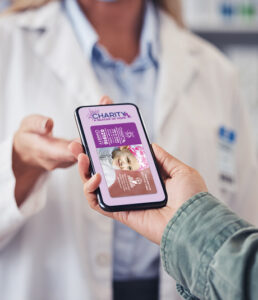 Image: a hand holding a cell phone with the CharityRx prescription discount card on the screen with a pharmacist in the background, reaching for the phone.
