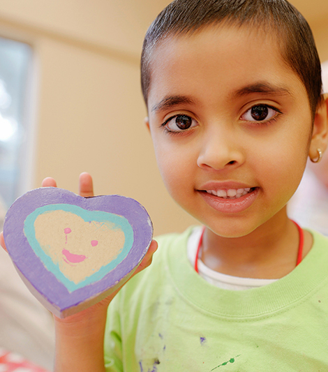 Image: a smiling, young Latina girl with very short hair holds a wooden heart that she painted.
