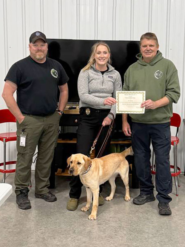 Image: Two middle-aged men wearing shirts with Jordan Detection logos stand on either side of a young blonde detective. In her left hand, she holds the leash of a young yellow lab, standing alertly. In her right hand, she holds one side of a certificate with the man on her left.