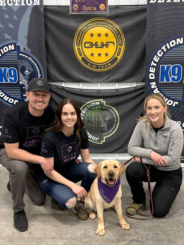 Image of a man wearing a CharityRx hat and t-shirt kneeling next to his wife, Charity, K9 Crusader and her handler, Shelby.