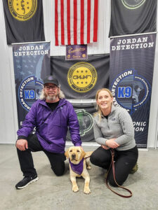 Image of a middle-aged man wearing a CharityRx hat and jacket kneeling next to Charity the K9 Crusader and her handler, Shelby.