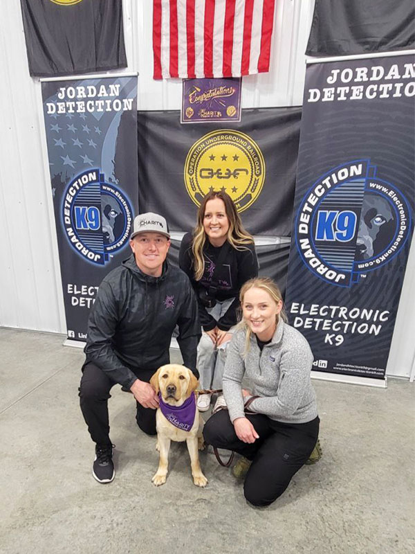 Image of a man wearing a CharityRx hat and jacket kneeling next to his wife, Charity the K9 Crusader and her handler, Shelby.