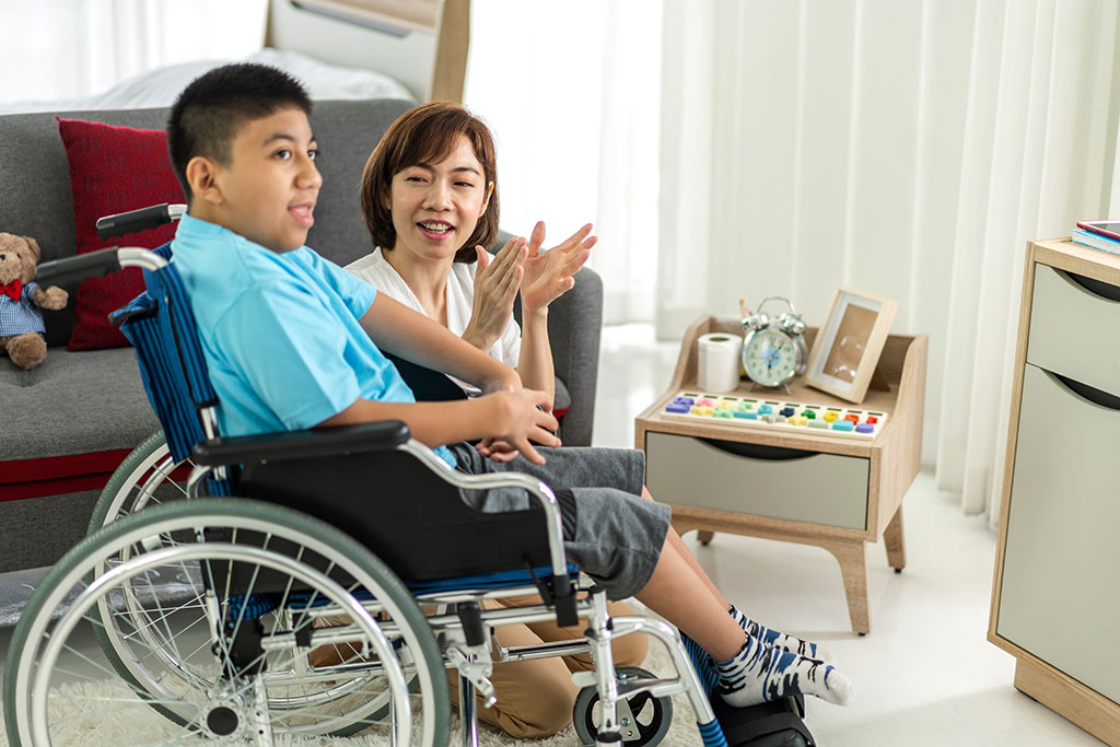 Image of a young, disabled Latinx boy sitting in a wheelchair next to an Asian woman who is kneeling next to the chair, singing and clapping her hands.